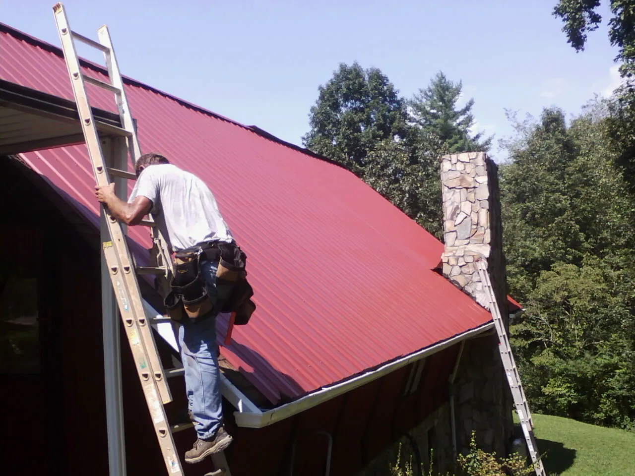Roofer on a ladder beside a red metal roof and stone chimney — Soffit & Fascia Repair by Roofing Brothers in Lakeville